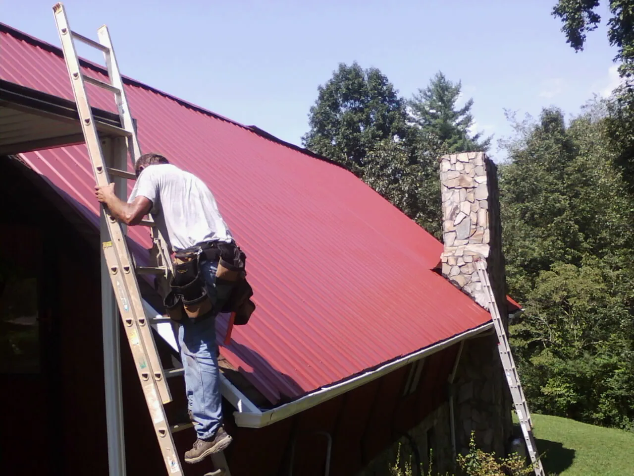 Roofer on a ladder beside a red metal roof and stone chimney — Soffit & Fascia Repair by Roofing Brothers in Waterford